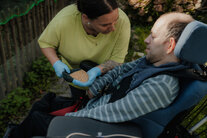 Eine Frau zeigt einem Mann im Rollstuhl eine Schaufel voll Getreide/Hühnerfutter
